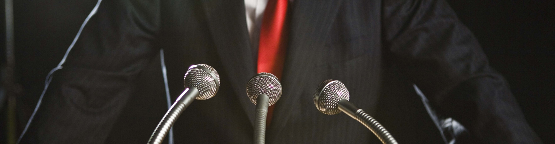 Close-up of a person in a dark suit and red tie standing behind three microphones